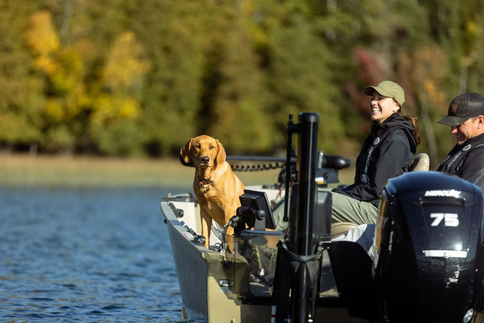 Dog on boat with two people on a lake, near autumn trees