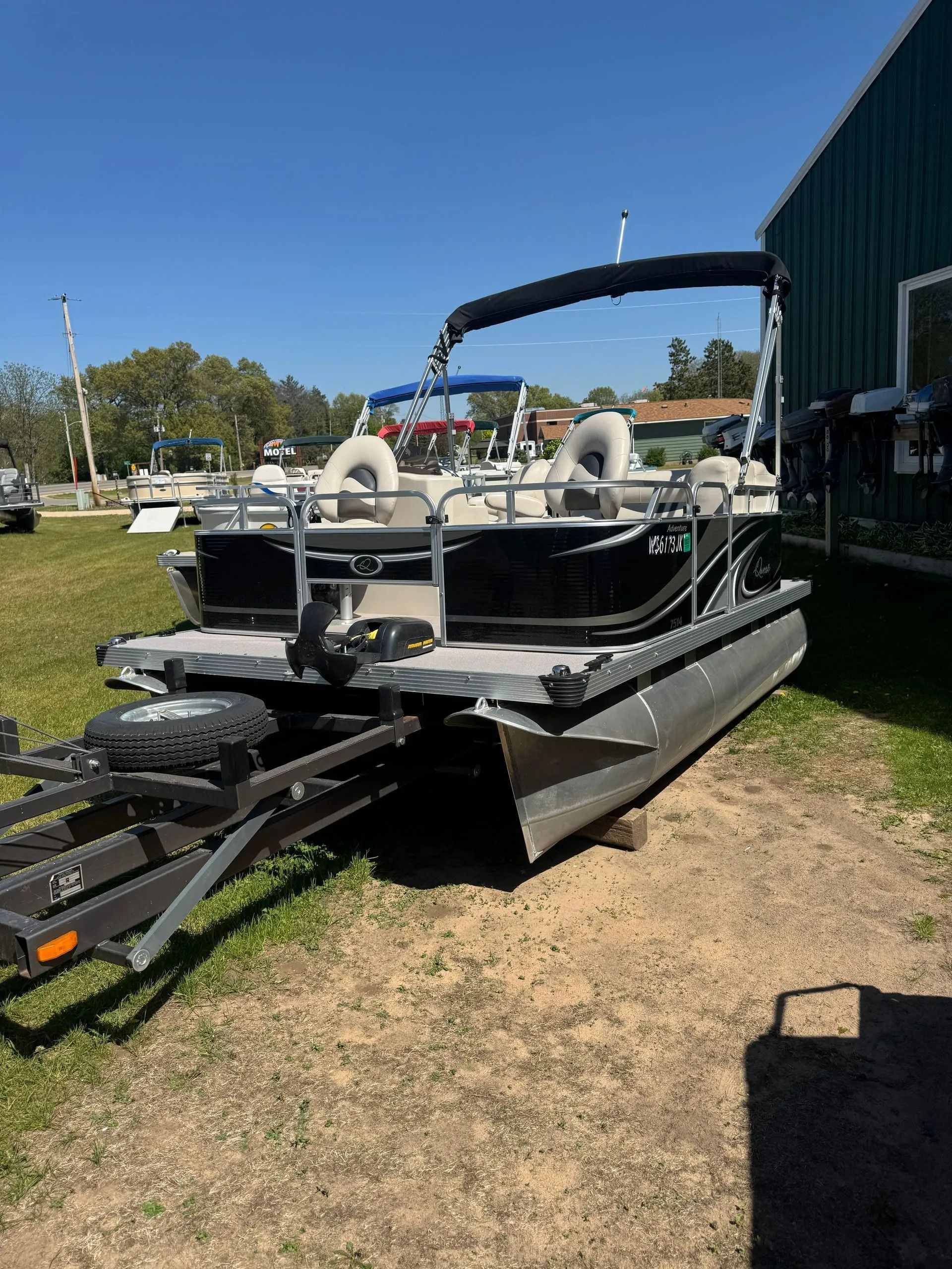 Pontoon boat on a trailer beside a building, with a blue canopy and white seats in a grassy lot