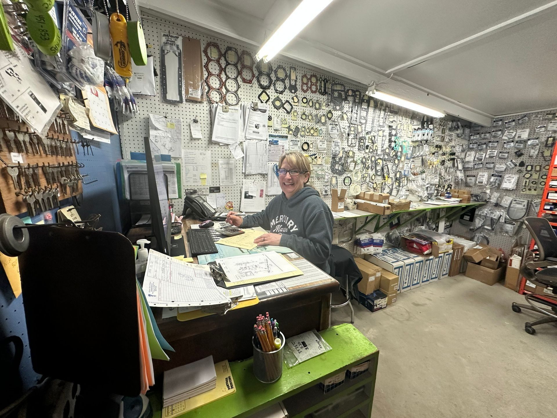 Person at a cluttered counter in a workshop, surrounded by tools, papers, and shelves of parts.