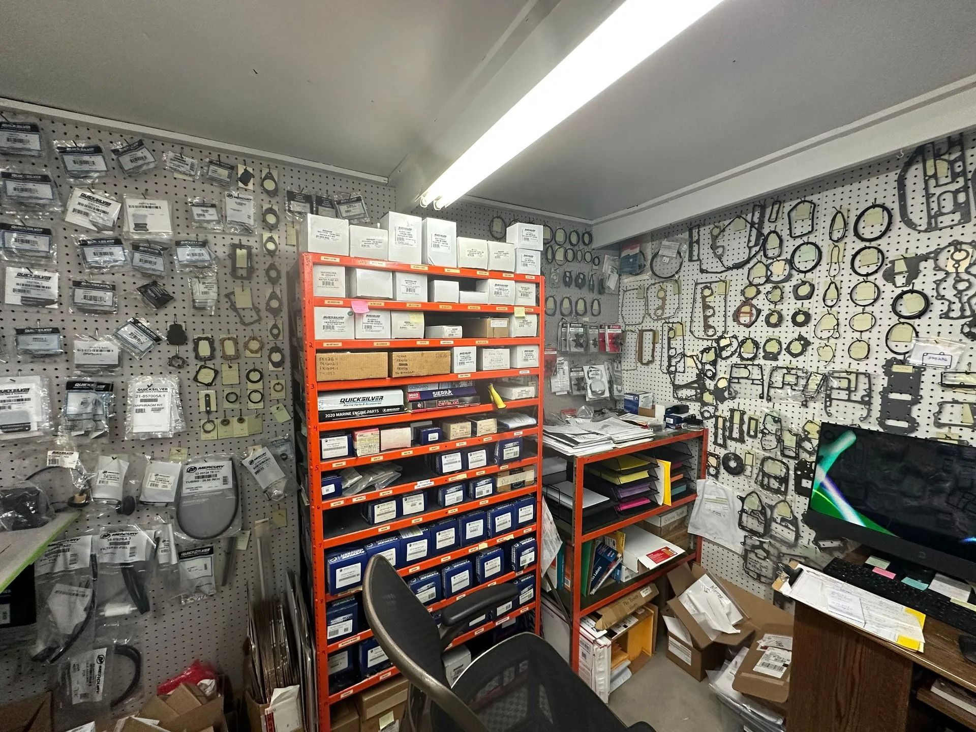 Cluttered workshop with shelves of small parts, wall-mounted tools, and a monitor on a desk.