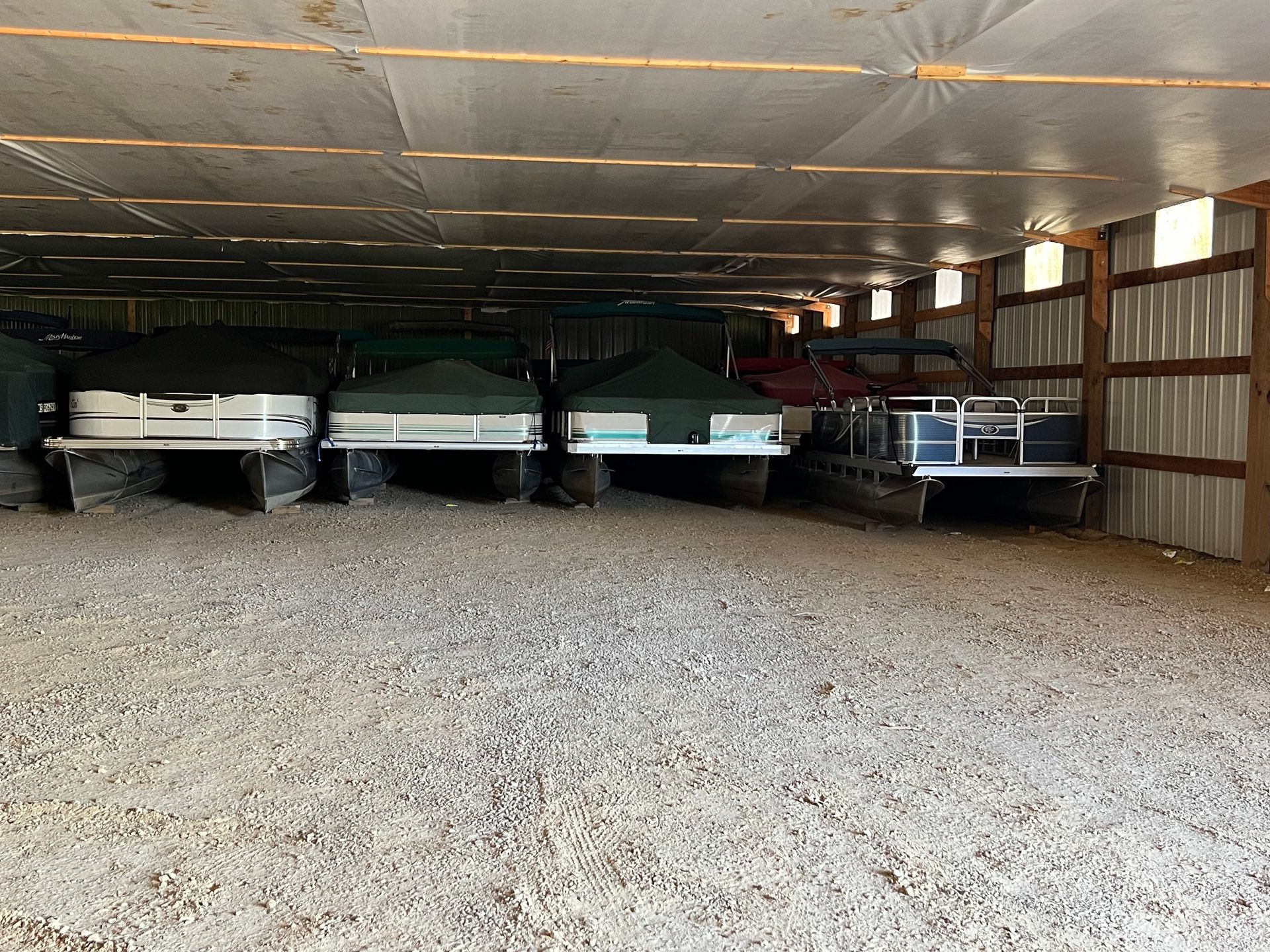 Inside a barn, several boats are stored on gravel under a metal roof with open sides.