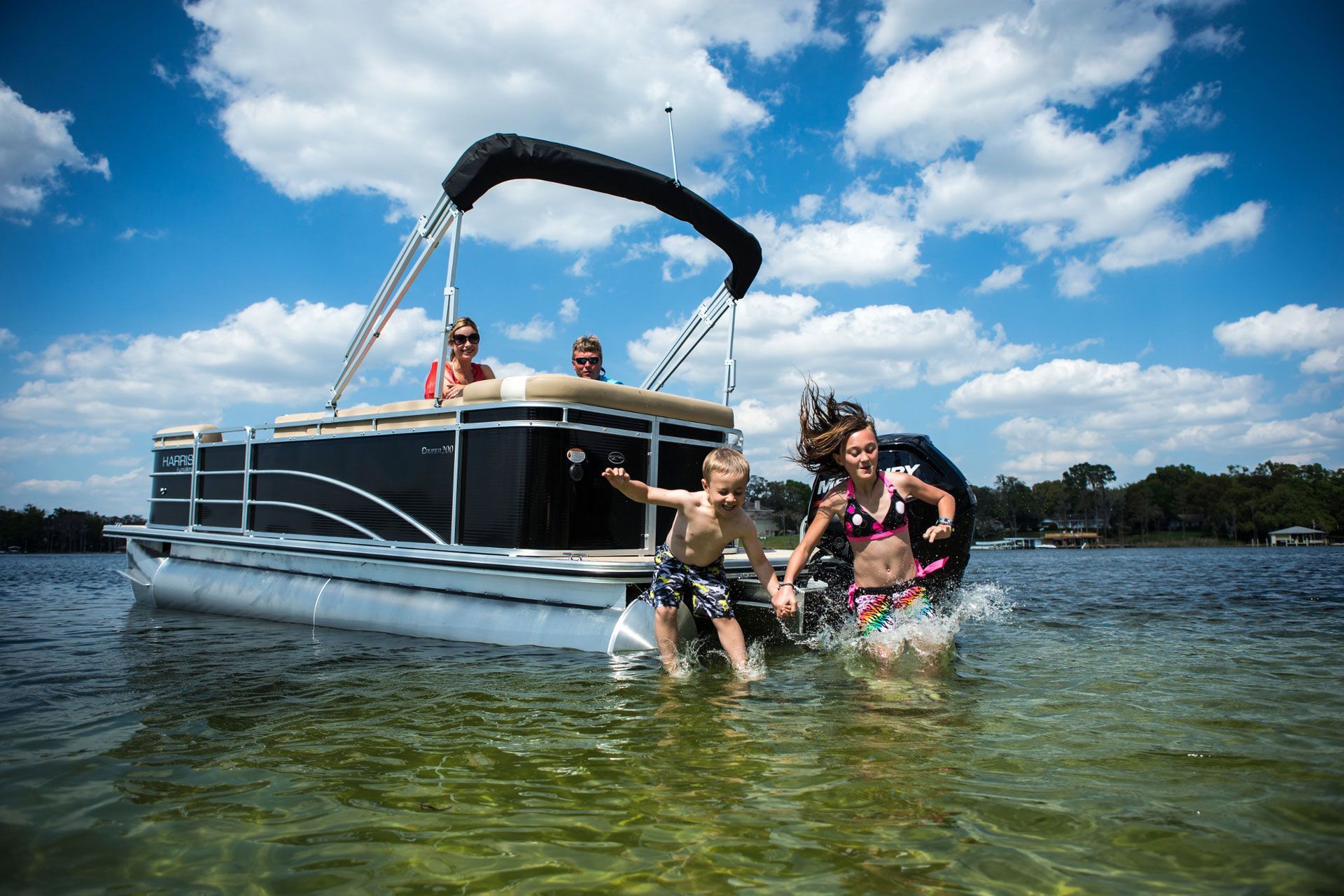 People swimming beside a pontoon boat on a sunny lake with blue sky and clouds.