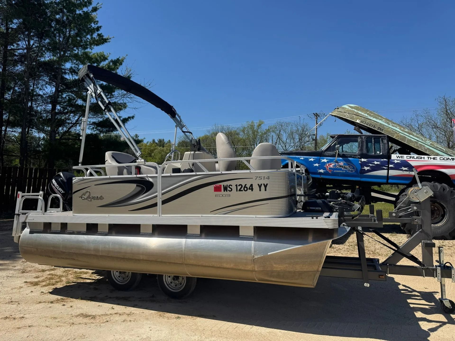 Pontoon boat on a trailer at a sunny outdoor lot, with trees and other boats in the background