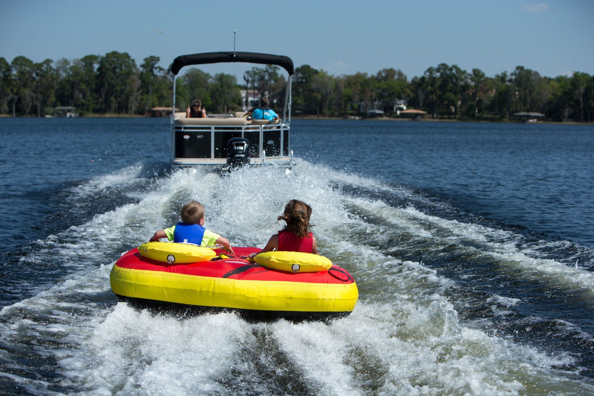 Two people ride a yellow-and-red towable tube behind a boat on a lake, splashing water.