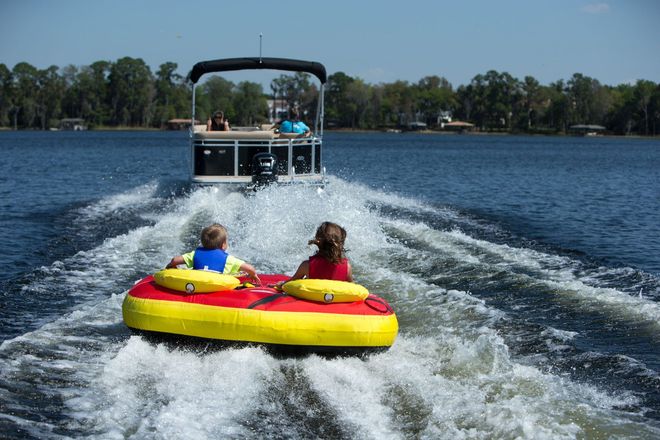 Two people ride a yellow-and-red towable tube behind a boat on a lake, splashing water.