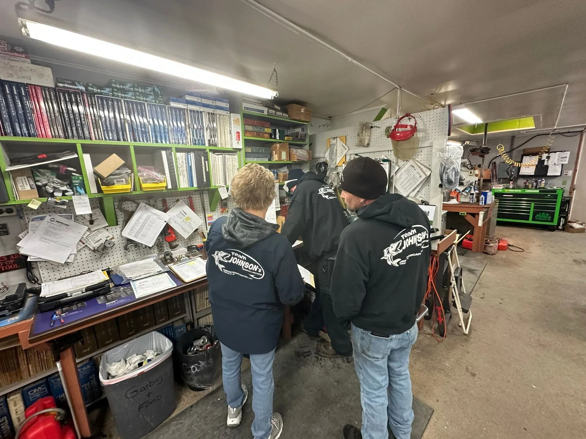 People in hoodies in a cluttered workshop, looking at papers on a wall of shelves.