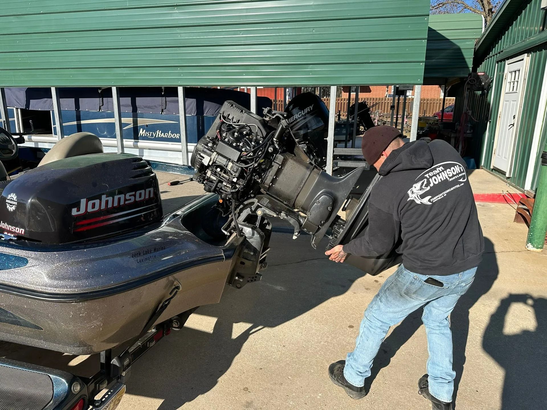Person loading a large Johnson outboard motor onto a boat trailer beside a green building