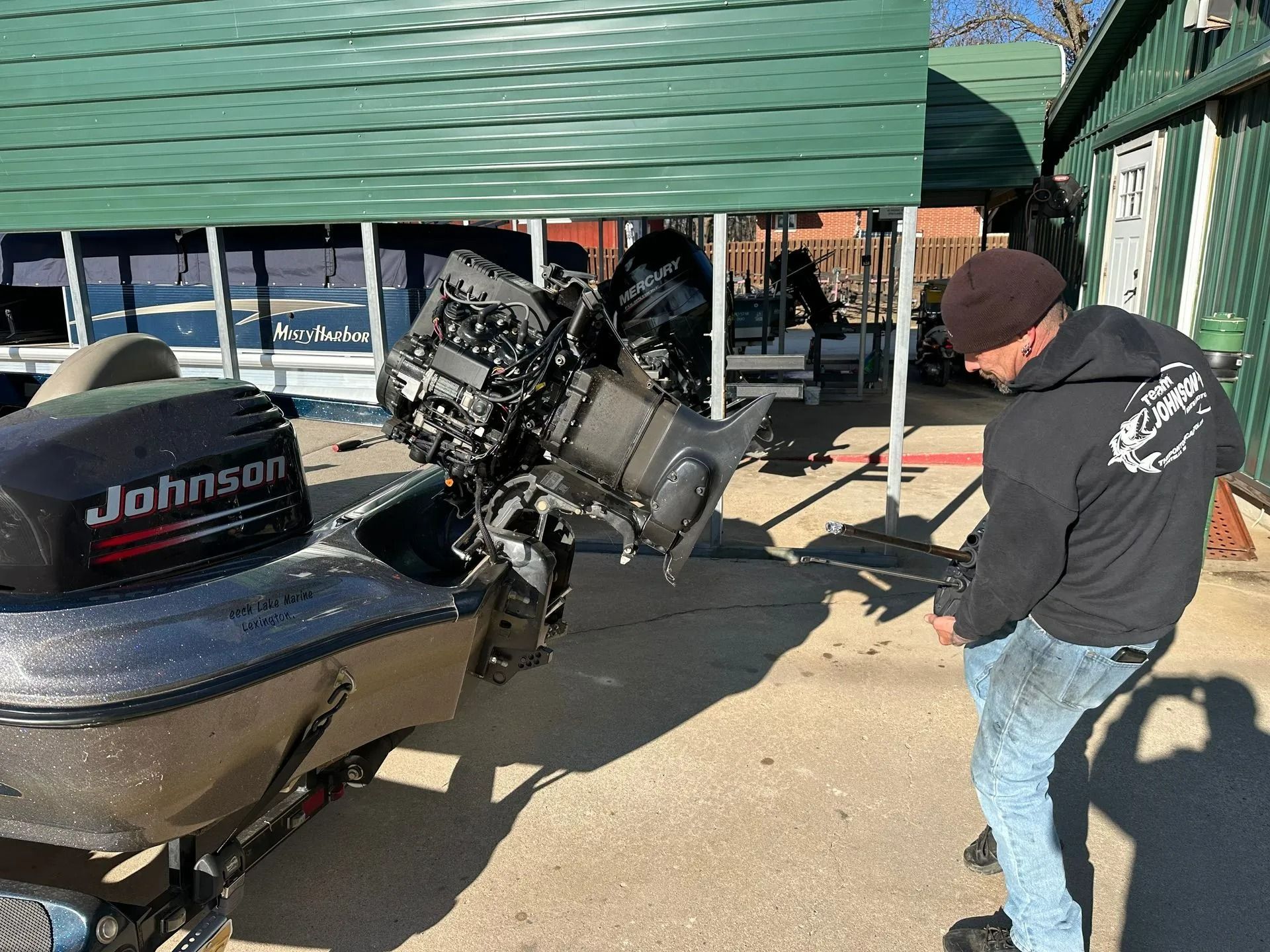 Man in black hoodie inspecting a boat’s outboard motor under a green canopy at a dock