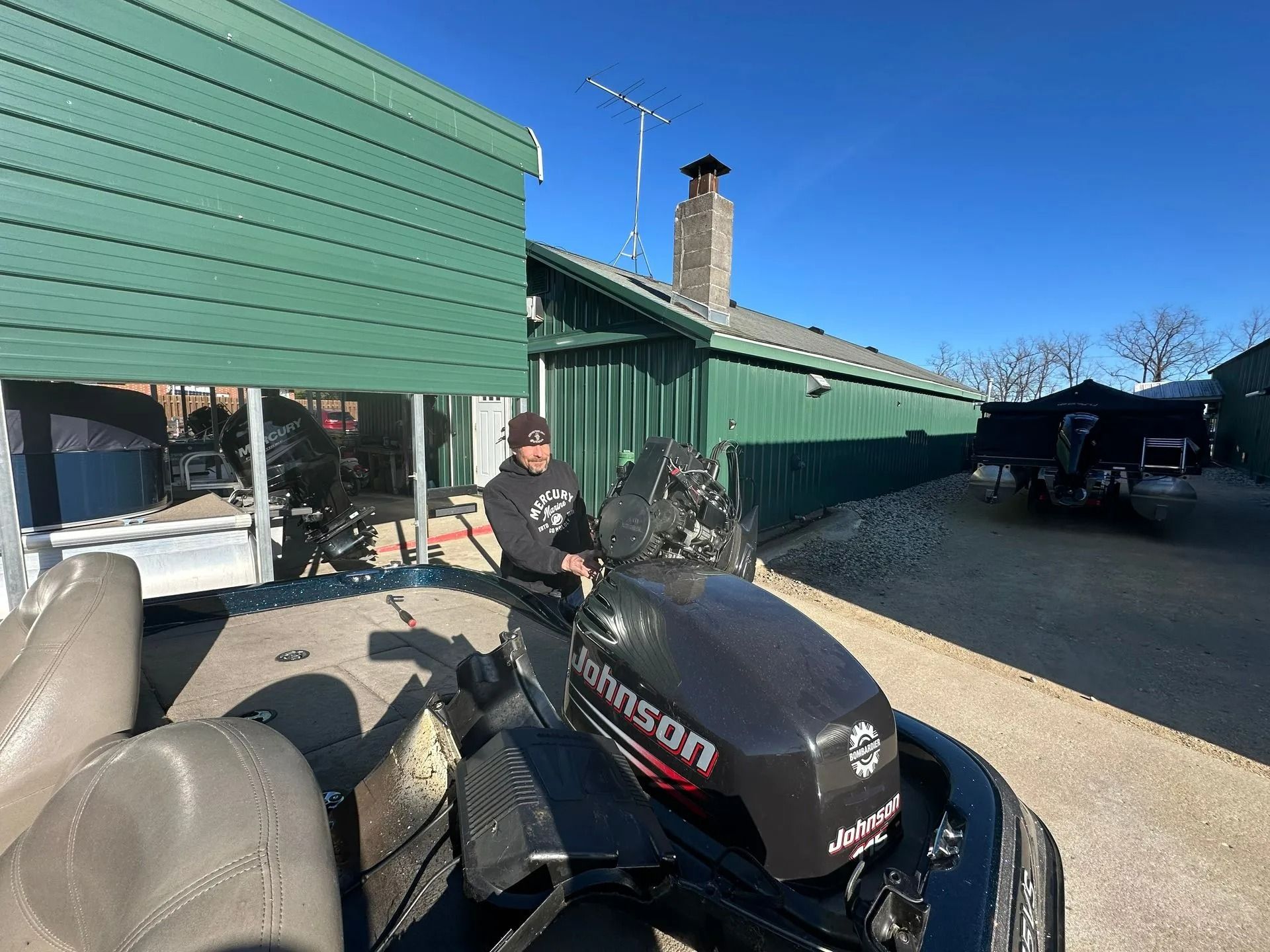 Person standing beside a black snowmobile in a driveway next to a green house under a blue sky