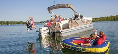 People boating on a lake, with one person jumping into the water near a pontoon and an inflatable tube.