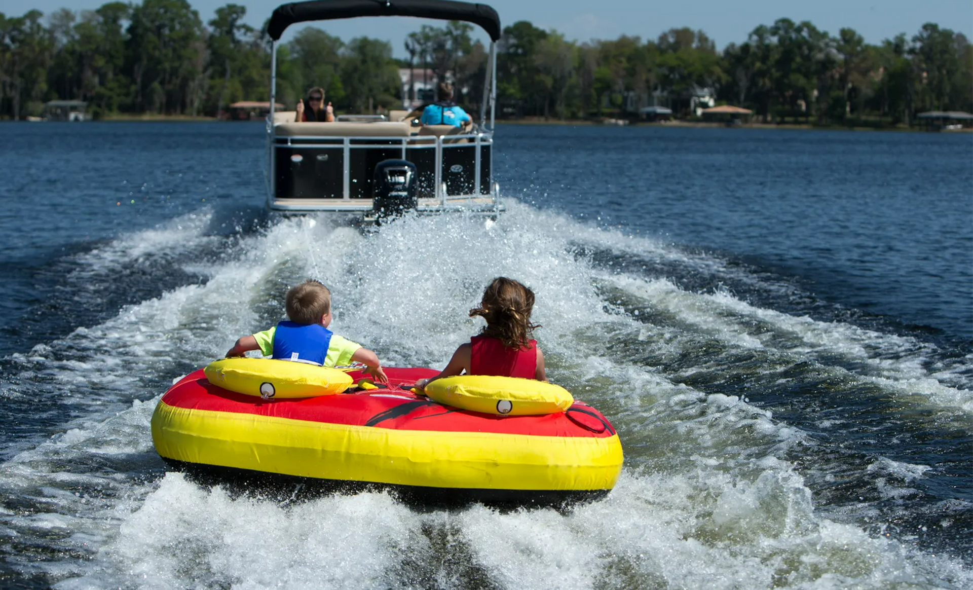 Two people on a yellow-and-red towable tube being pulled behind a boat on a lake