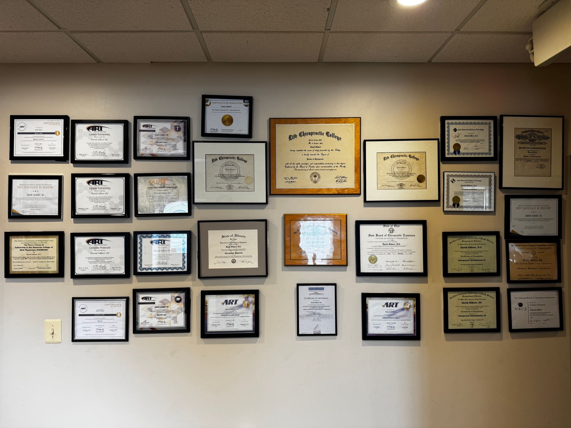 A wall covered with numerous framed diplomas, certificates, and awards arranged in a grid-like display.