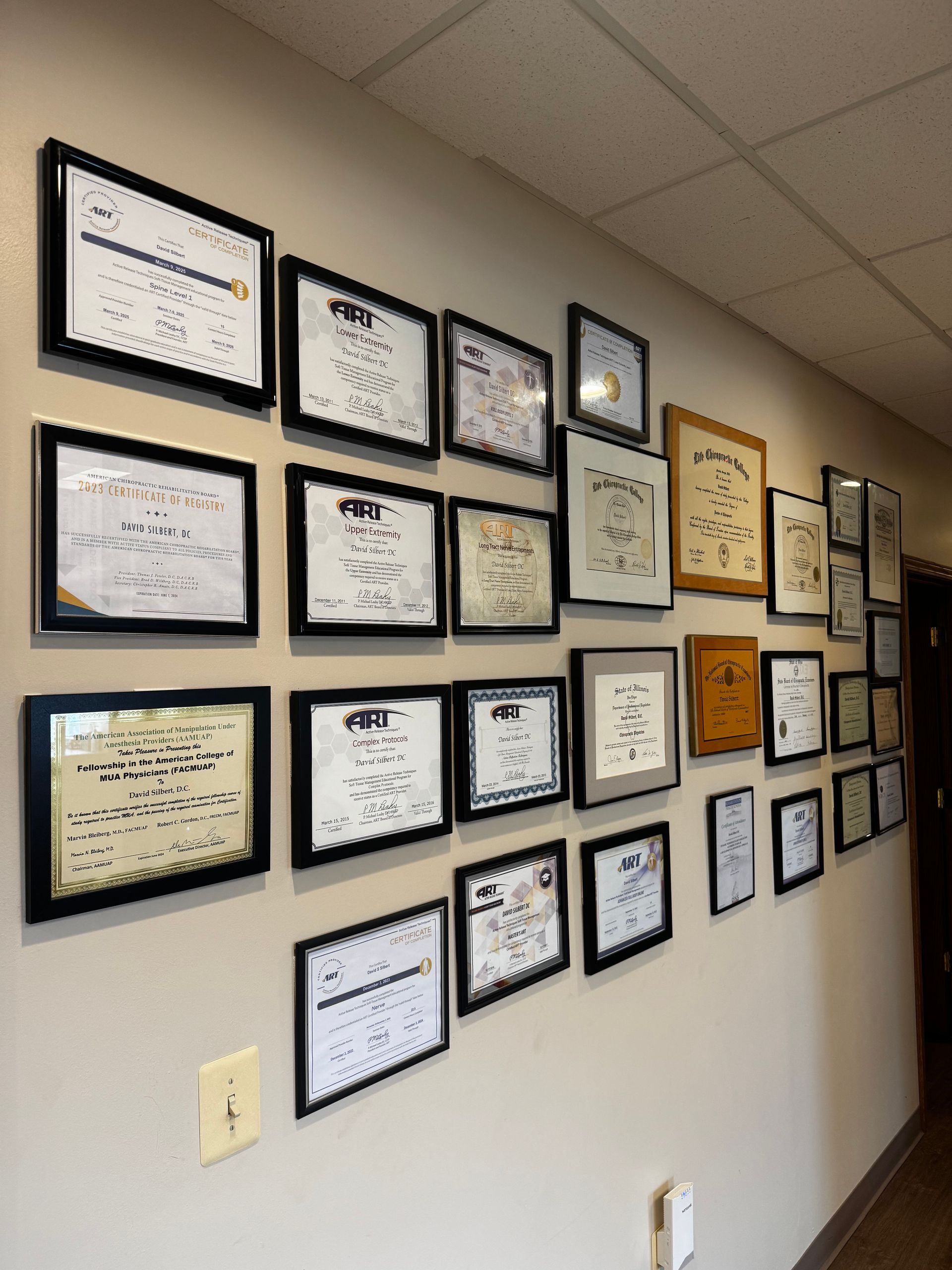 A wall in a professional office displaying many framed certificates and diplomas of various sizes.