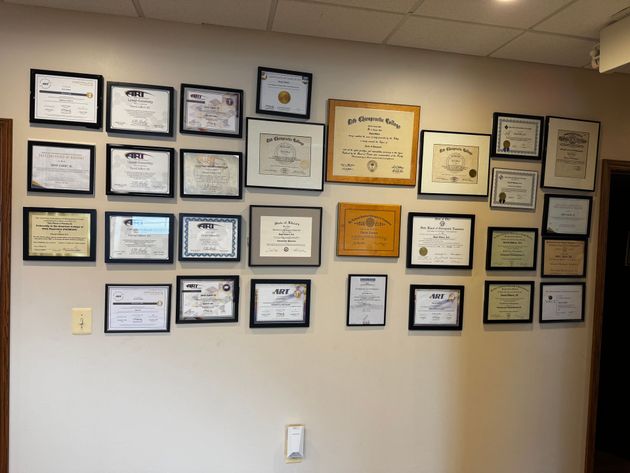 A wall covered in numerous framed certificates, diplomas, and awards arranged in a grid-like display in an office setting.