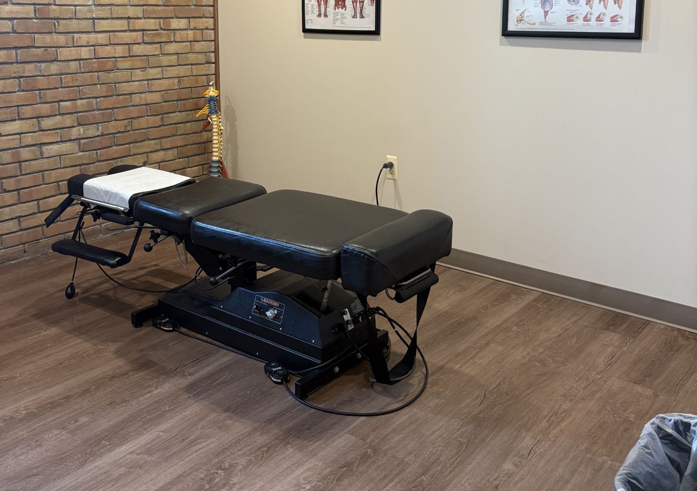 Chiropractic treatment table in a small exam room with brick accent wall and framed charts.