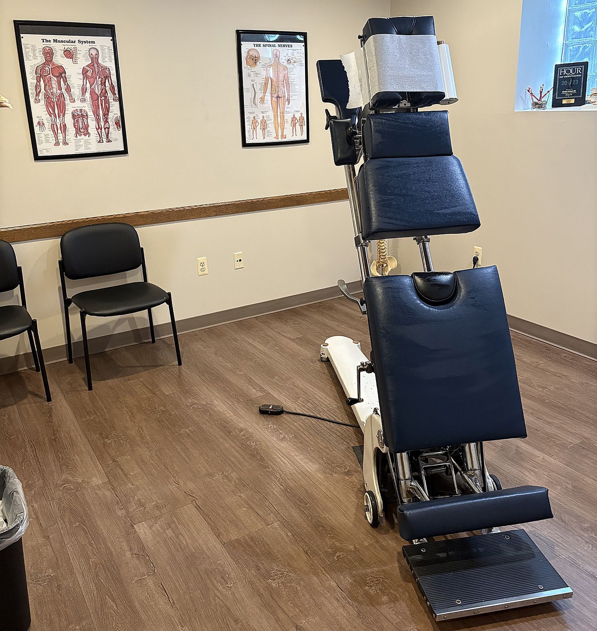 Chiropractic treatment room with an adjustable exam table, chair, anatomy posters, and wood flooring.