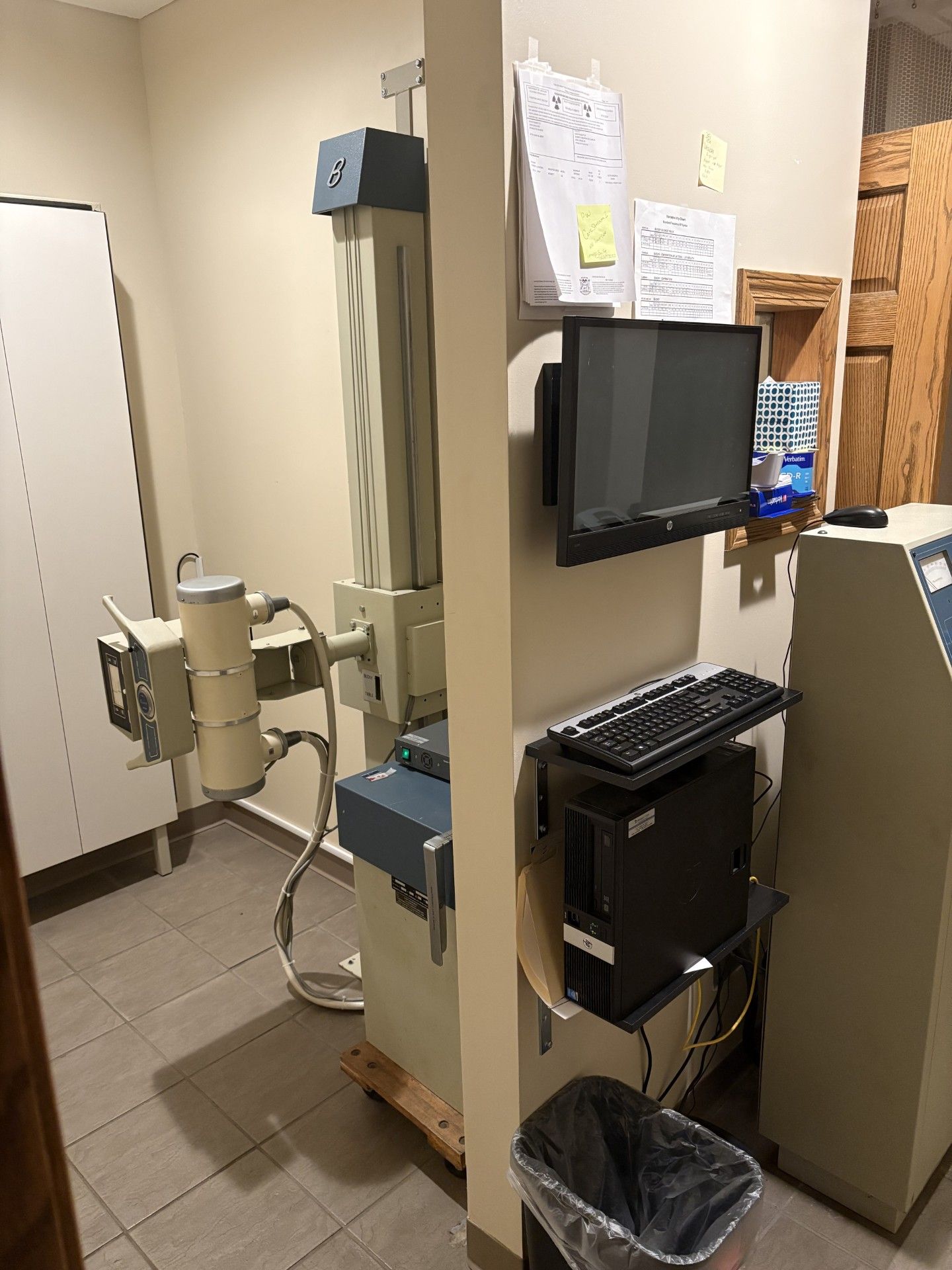 Clinic room with dental x-ray machine, computer monitor, keyboard, and trash bin.