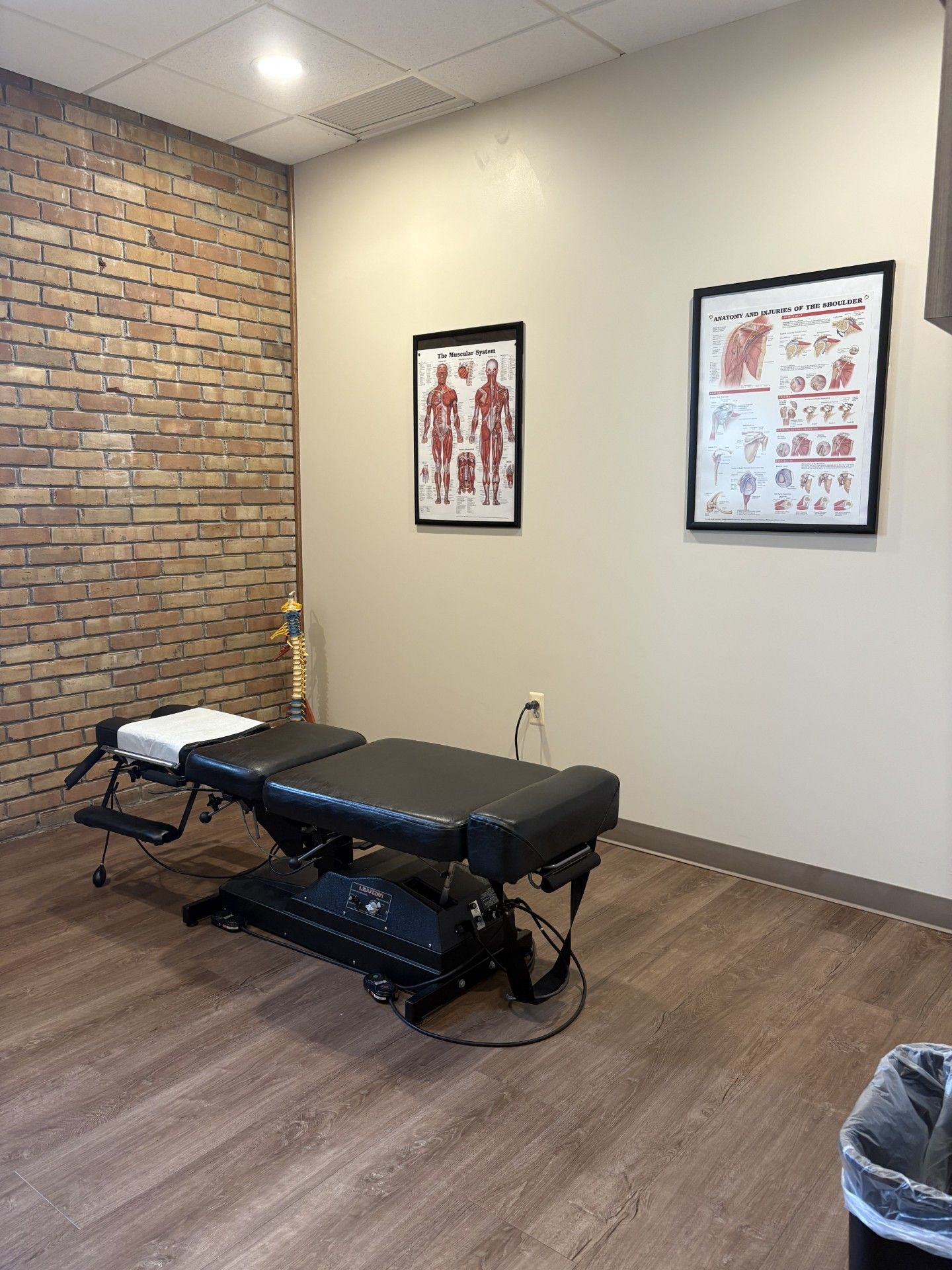 Empty chiropractic treatment room with an adjustable table, brick accent wall, and anatomical posters.