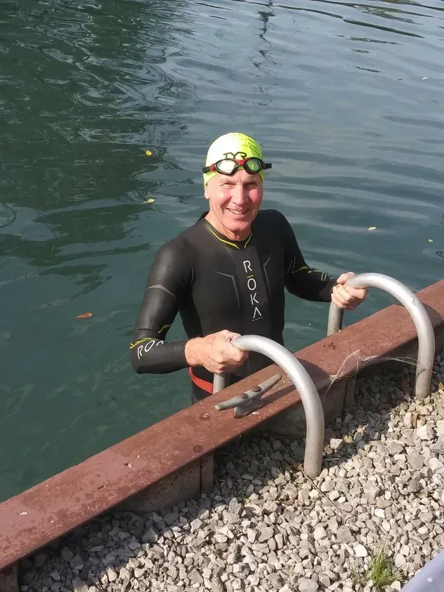 A smiling person in a wetsuit and neon cap holds onto a dock ladder after swimming in a lake.