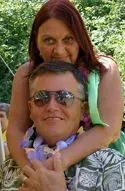 A person with red hair embraces someone wearing sunglasses and a patterned shirt, both smiling outdoors.