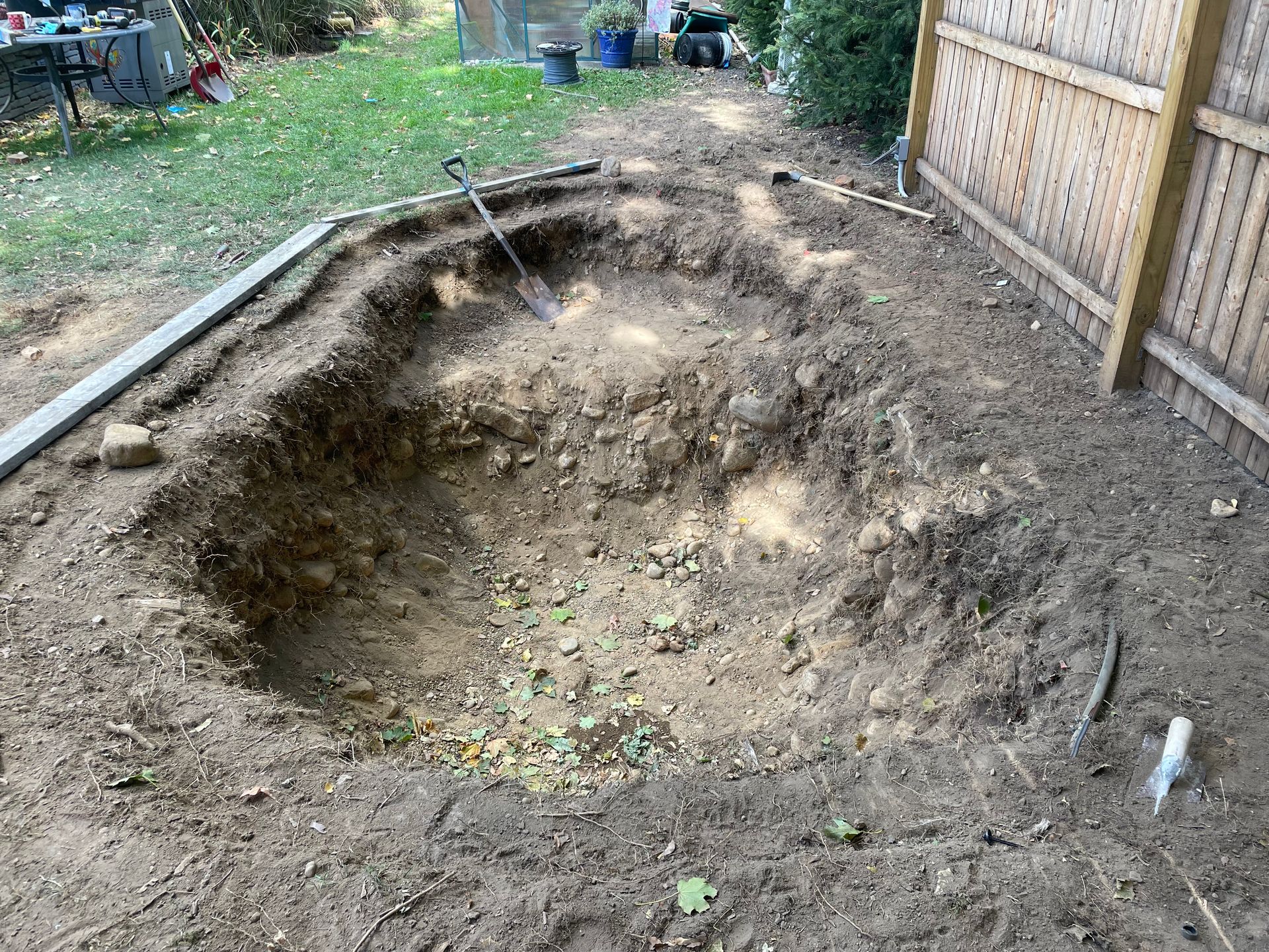 Large dug-out dirt pit in a backyard beside a wooden fence and garden area.