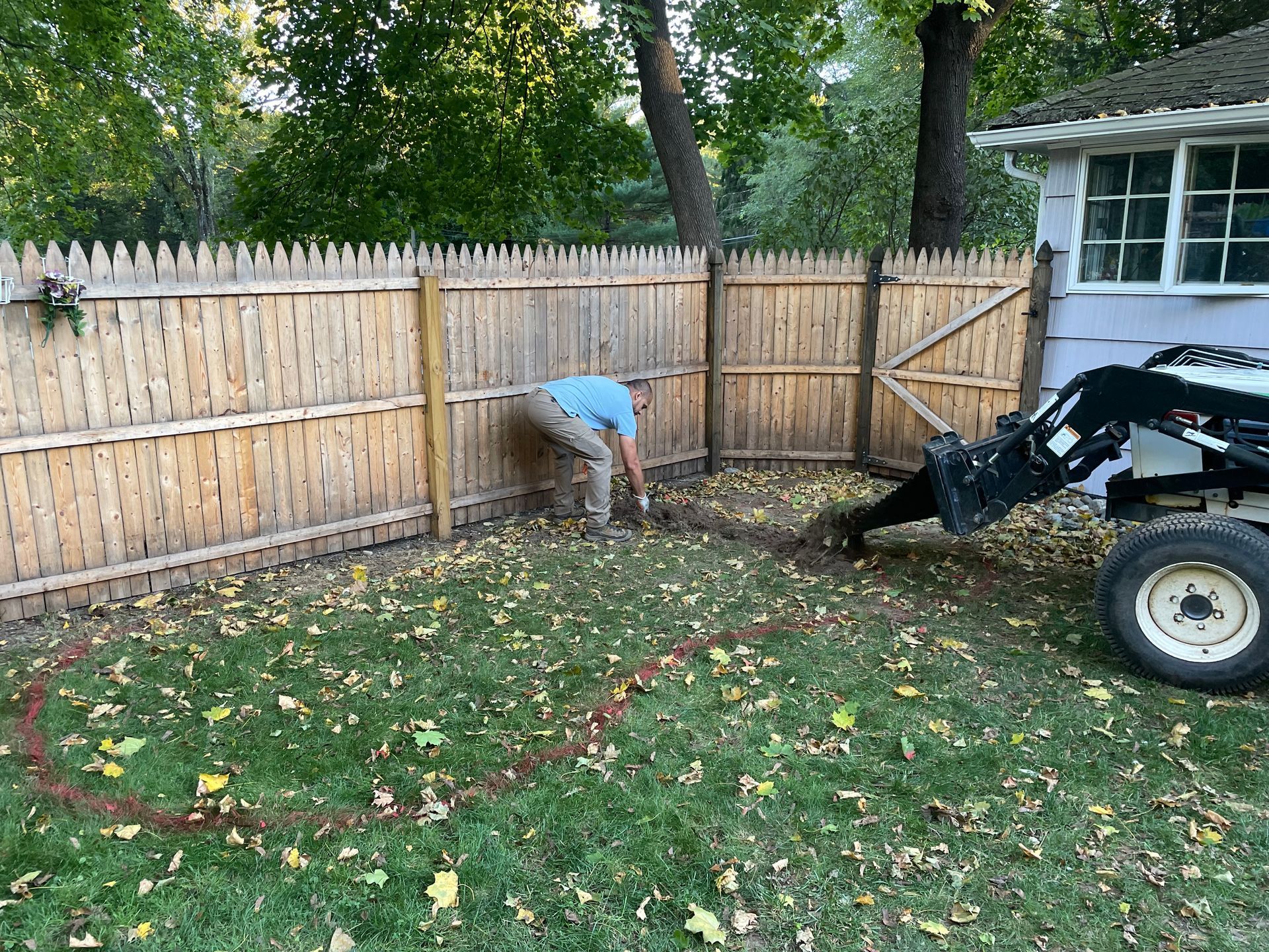 Person digging a backyard garden bed beside a wooden fence, with a small tractor nearby.
