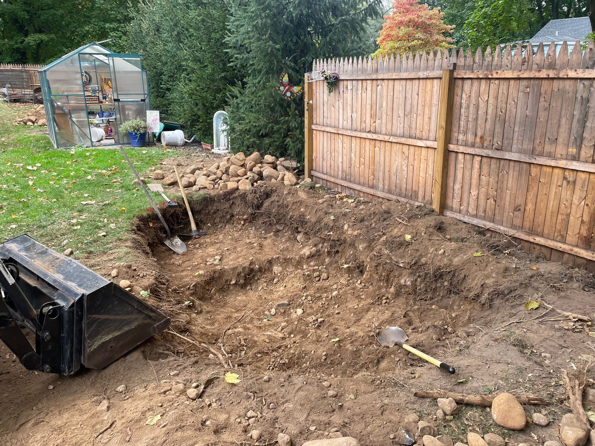 Backyard excavation with a large dirt pit, rocks, a shovel, and a wooden fence beside a shed