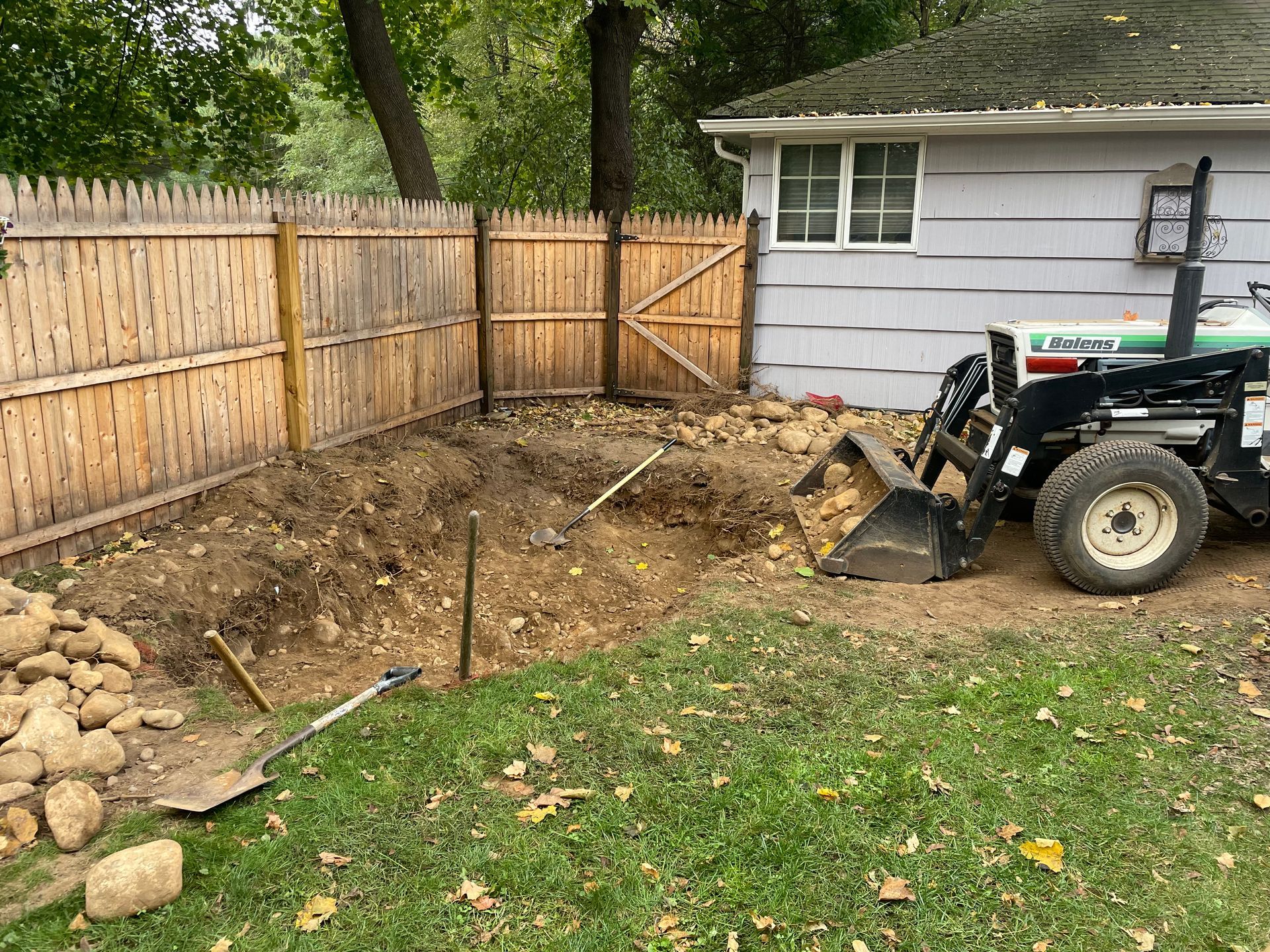 Backyard excavation beside a shed, with a tractor, wooden fence, and dirt pile.