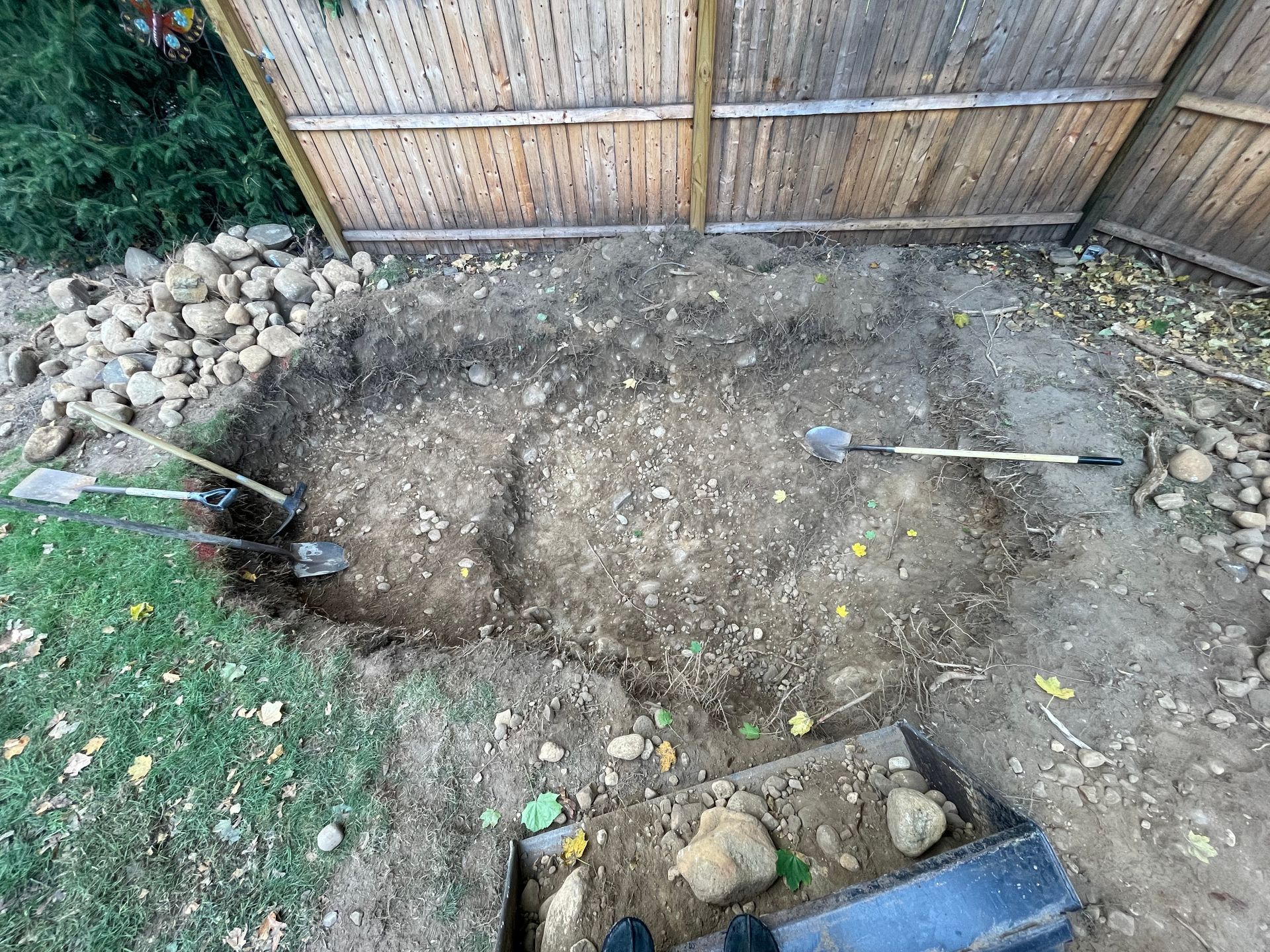 Bare dirt backyard area with a wooden fence, rocks along the edges, and garden tools on the ground