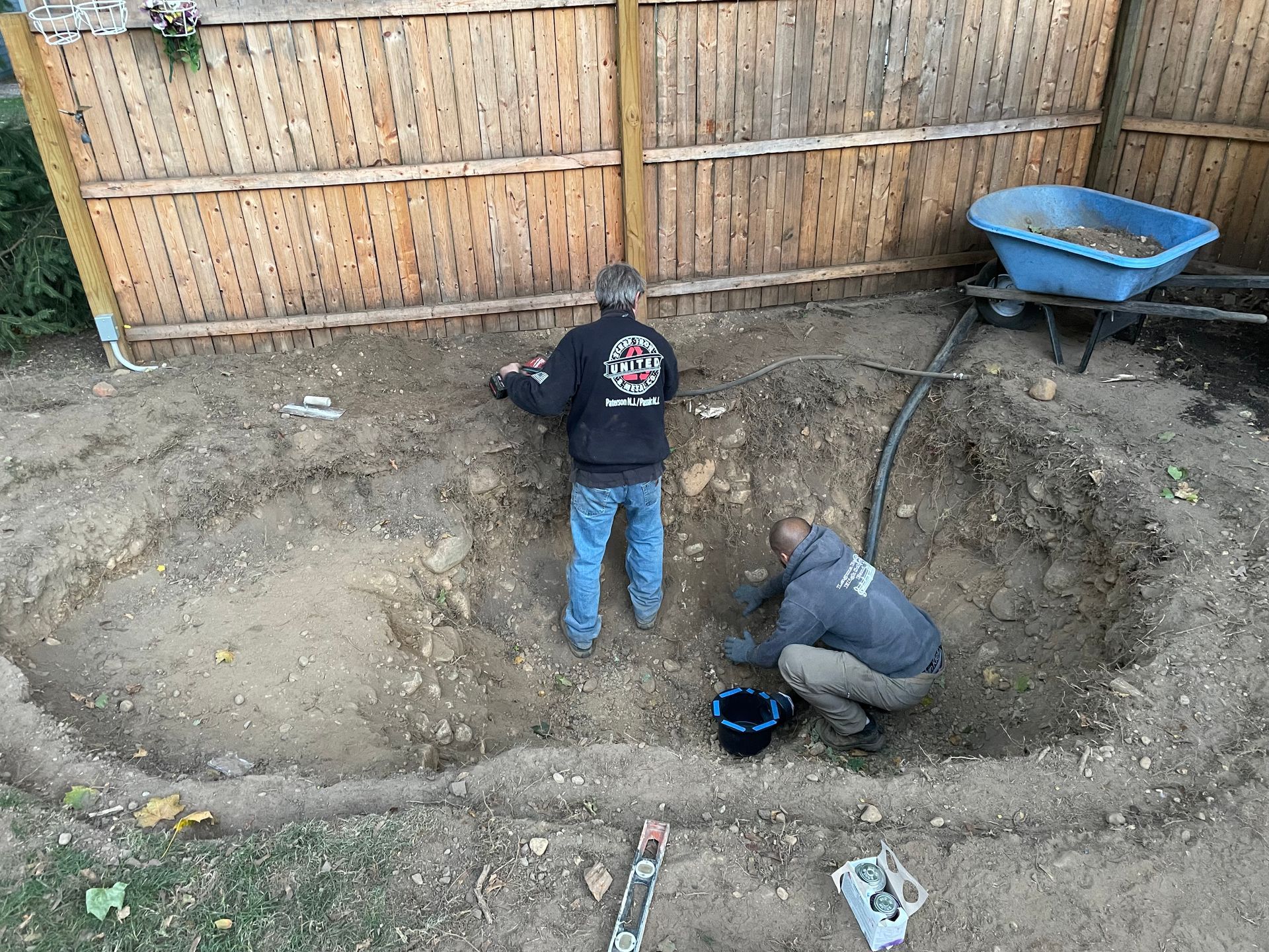 Two people digging a large backyard pit beside a wooden fence and blue wheelbarrow.