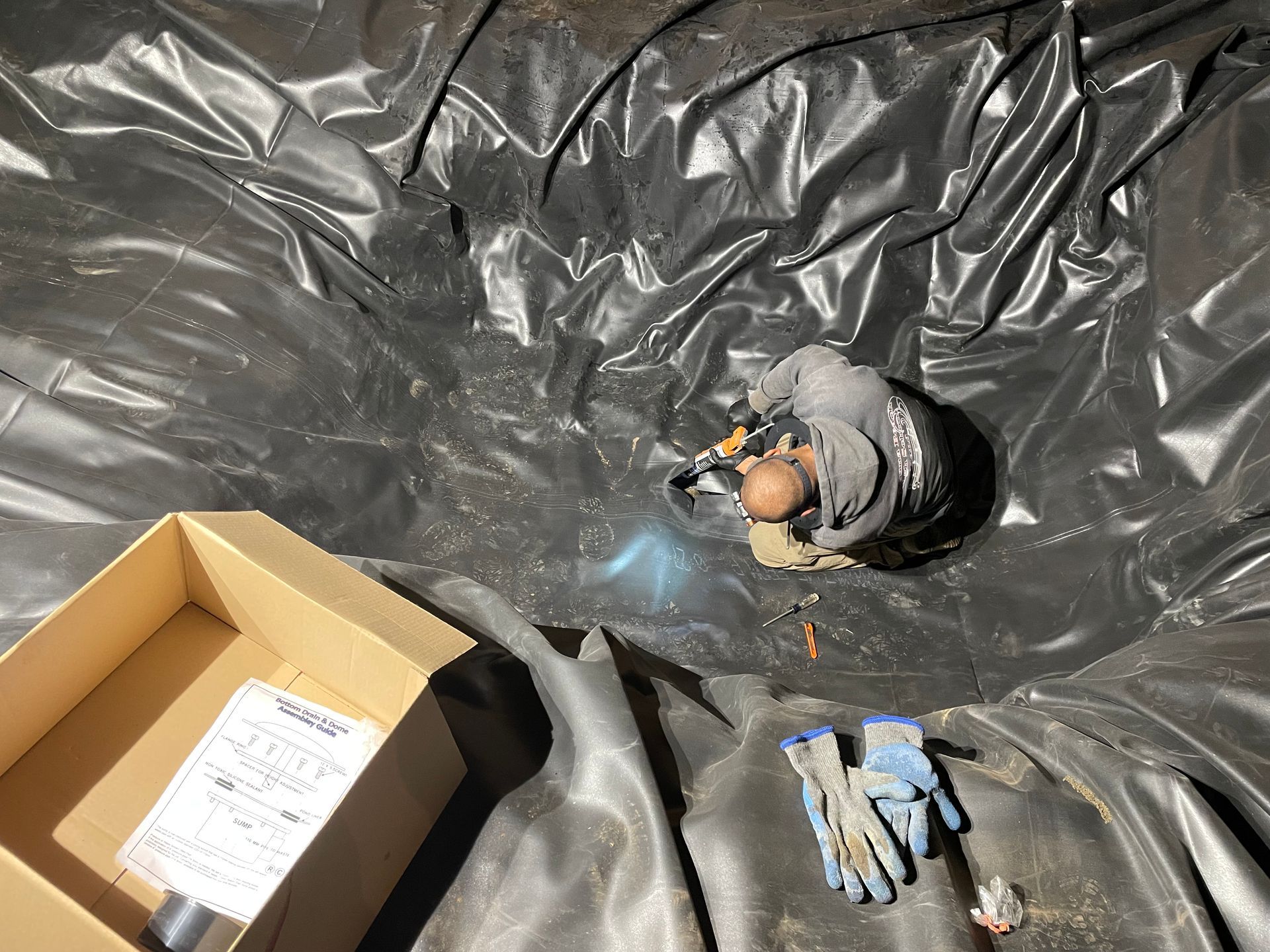 Person in a black plastic-lined pit, working with a tool near a box of papers and gloves.