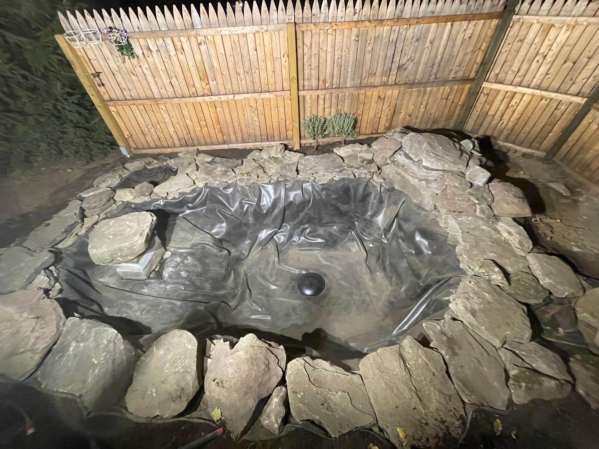 Stone-lined backyard pond under a bamboo fence, lined with black plastic and filled with water.