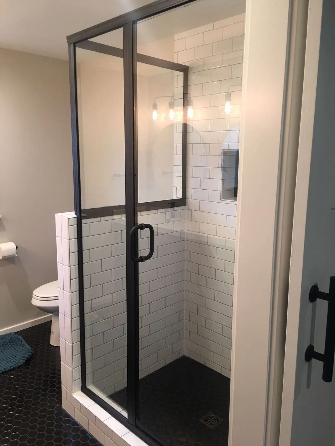 Black-framed glass shower with subway tile walls and black hexagon floor tiles. Toilet visible in the background.