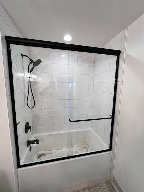 A black-framed glass shower door over a white bathtub in a bathroom with white tile walls.
