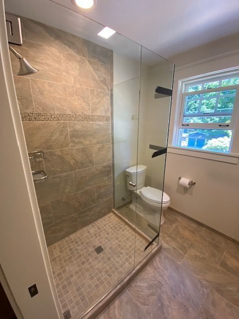 Bathroom with tiled shower, glass door, toilet, window, and neutral-toned flooring and walls.