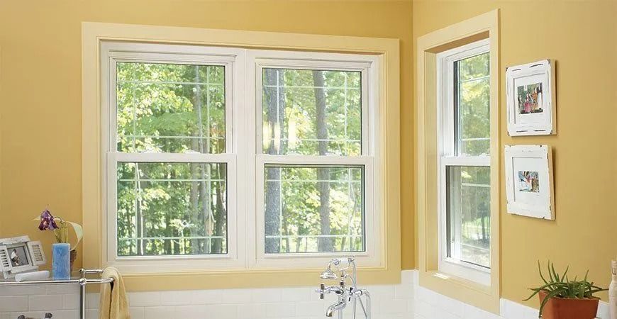 Yellow-walled bathroom with two white-framed windows overlooking a green tree line; two picture frames and a plant.