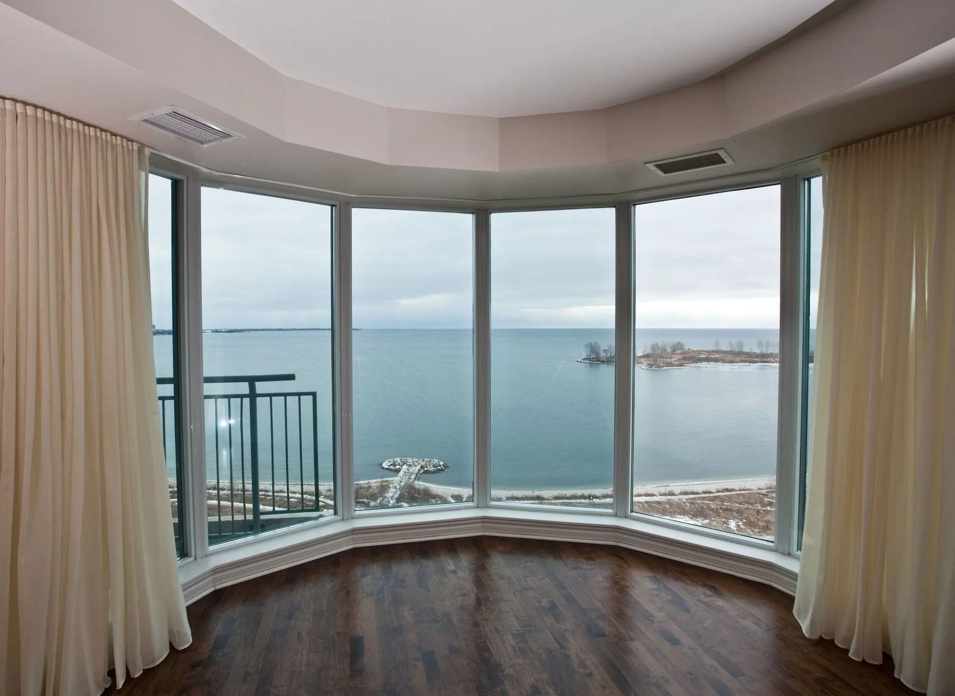 A bright room with curved windows overlooking a body of water and a city. Beige curtains frame the windows.