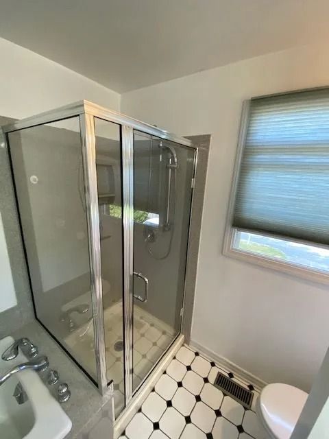 Shower stall with glass doors and silver trim in a bathroom. White walls, black and white tiled floor, and a window with a blind.