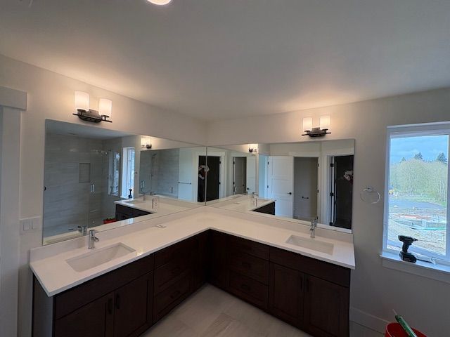 A modern bathroom vanity with dark wood cabinets, white countertops, two sinks, and an L-shaped mirror, under wall lighting.
