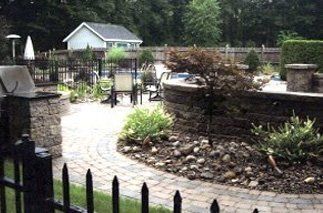 Outdoor patio with a pool, grill, and landscaped yard behind a black wrought iron fence.