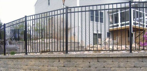 Black metal fence atop a retaining wall, enclosing a house with a deck.