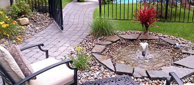 Stone pathway leads to a patio with a small fountain surrounded by plants and a red plant near a black fence.