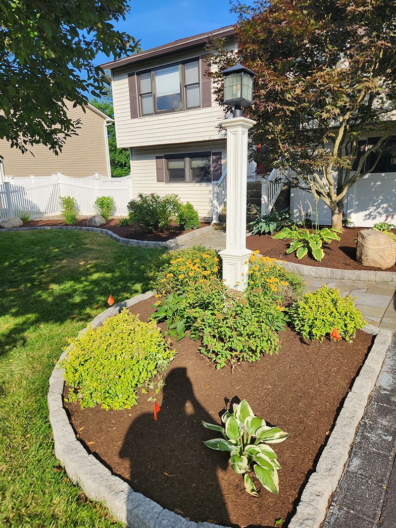 A house with landscaping, a lamp post in a garden bed with flowers and mulch. Sunny day.