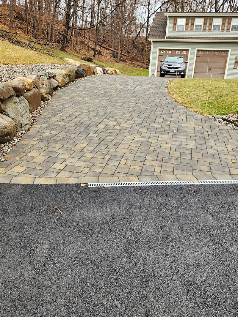 Brick driveway leading to a garage.  Driveway is light brown and tan. Garage has brown doors.  Car parked inside.