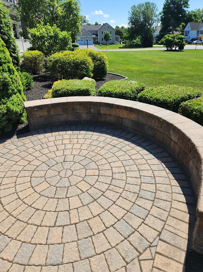 Circular brick patio with a low stone wall, surrounded by green shrubs and grass lawn.