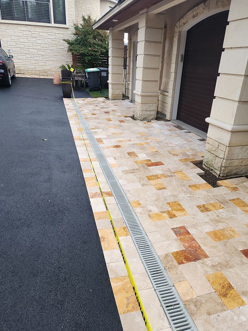 Asphalt driveway next to stone walkway with a drainage grate near a garage.