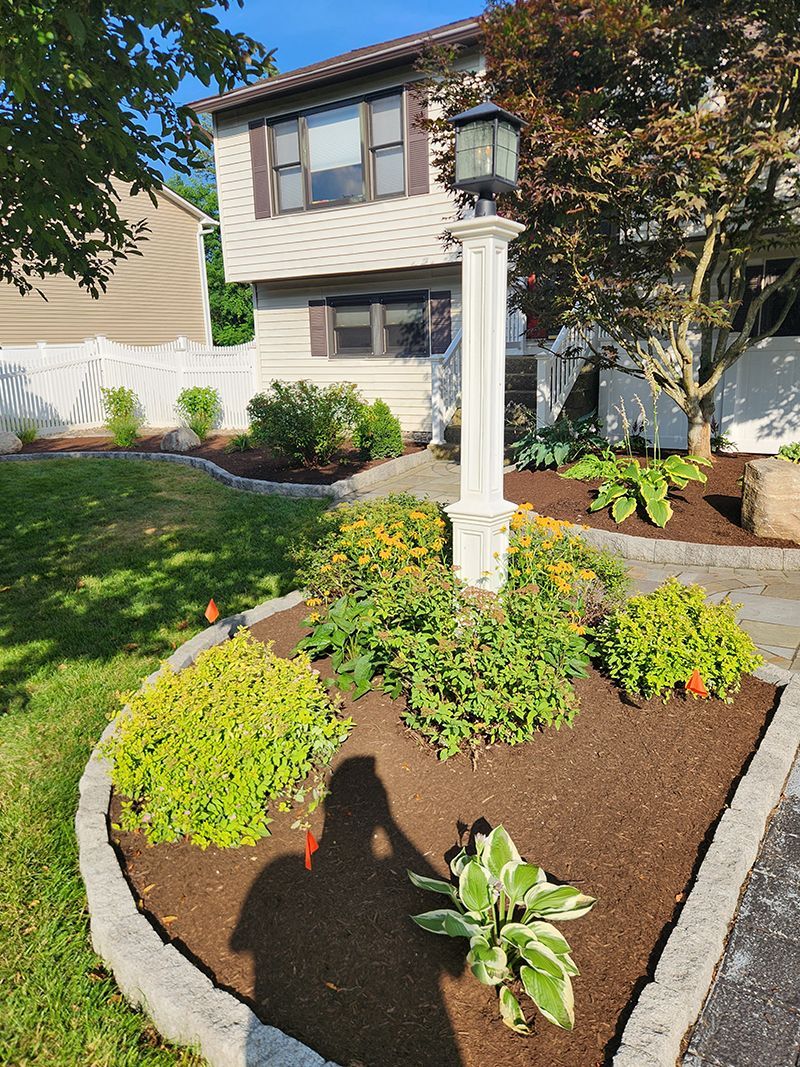 Lawn with flower beds, a white lamppost, and a two-story house. The sun is shining.