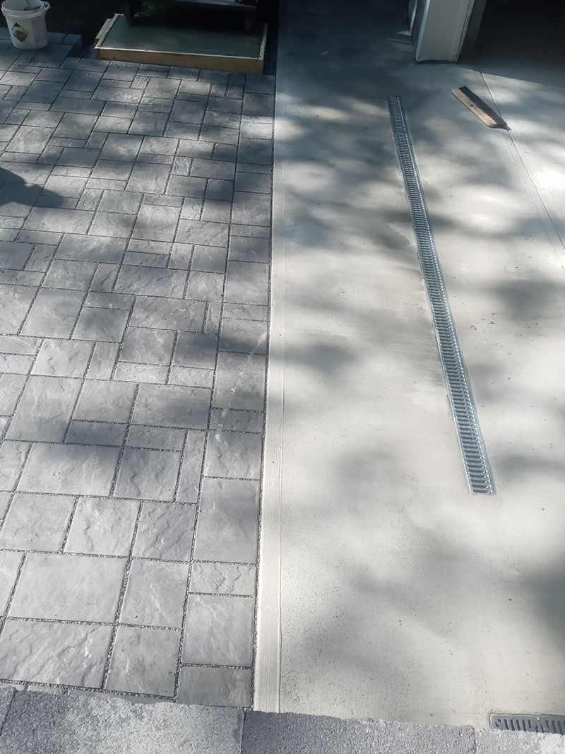 Gray paver walkway next to a concrete surface with a drain.