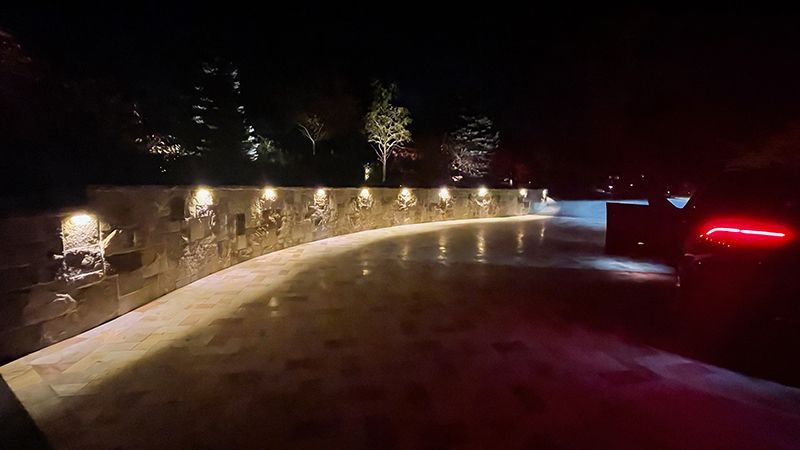 Curving, illuminated driveway at night, with a retaining wall and car taillights visible.