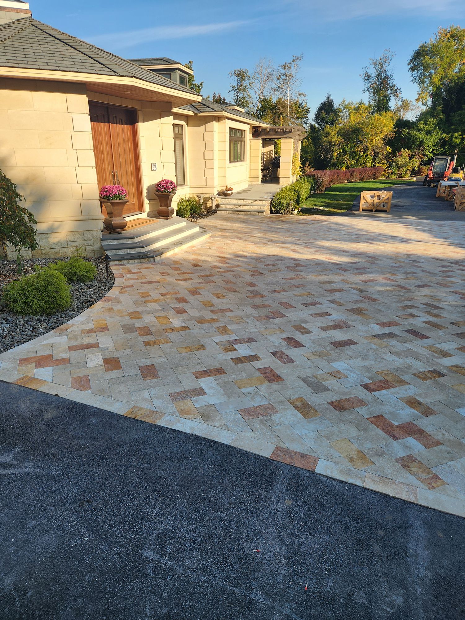 Brick-paved driveway leading to a light-colored stone house with a brown door. Landscaped front yard with green lawn.